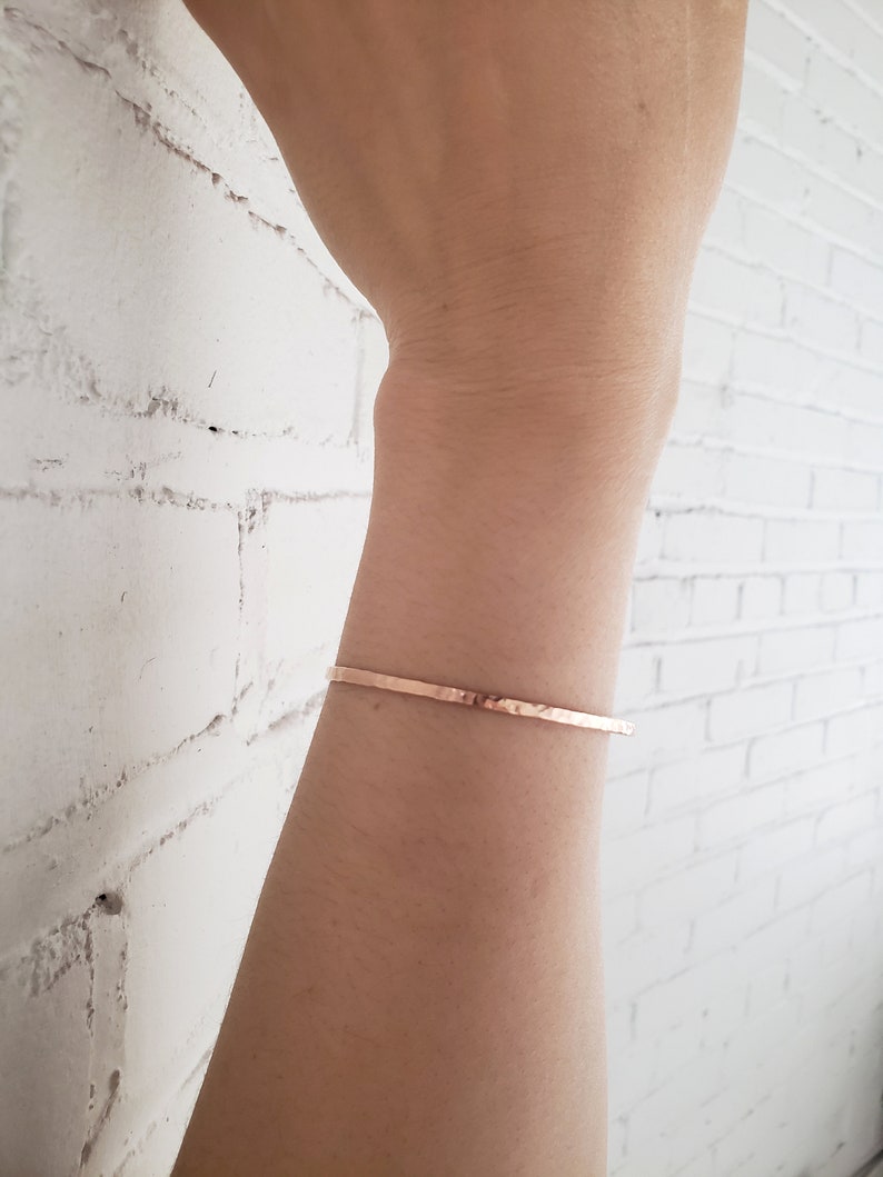 May include: A thin, hammered, rose gold bracelet on a person's wrist.