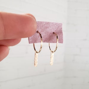 Puede incluir: Aretes de aro de oro con un colgante de barra martillada. Los aretes est&aacute;n sobre un fondo rosa.