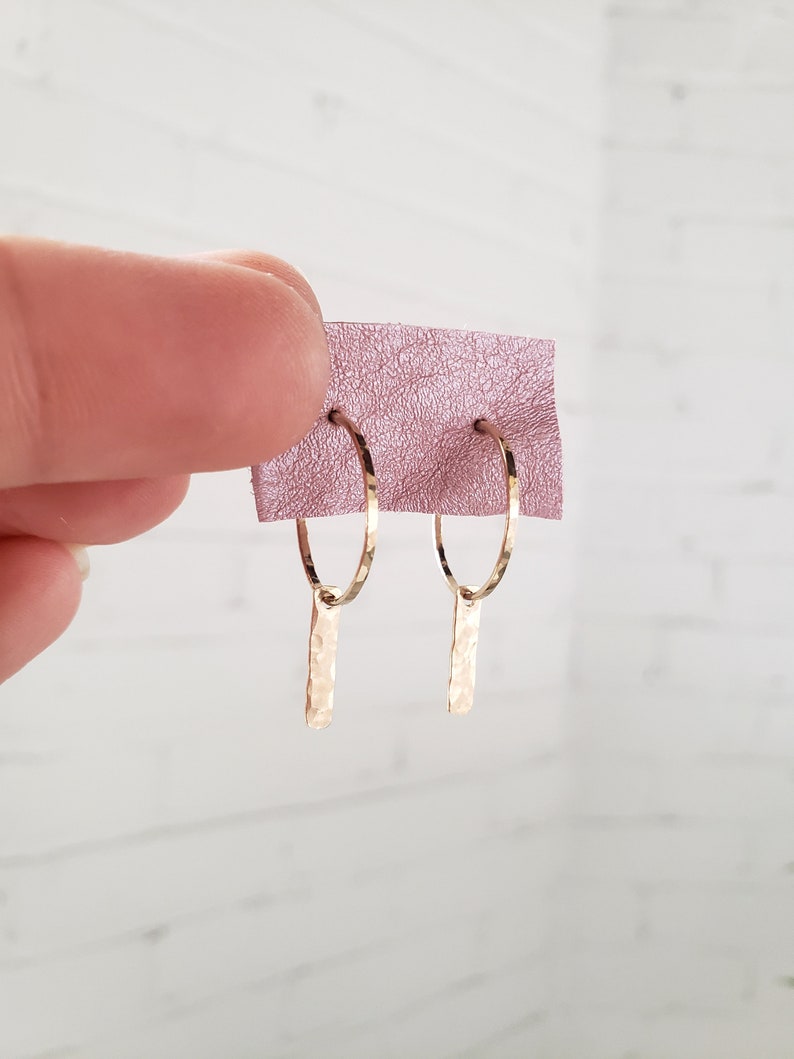 Puede incluir: Aretes de aro de oro con un colgante de barra martillada. Los aretes est&aacute;n sobre un fondo rosa.