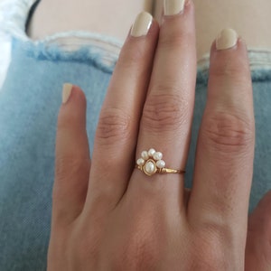 Peut inclure: Une bague en or avec un groupe de petites perles blanches en forme de fleur. La bague est sur le doigt d'une personne.
