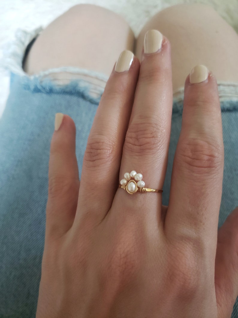 Peut inclure: Une bague en or avec un groupe de petites perles blanches en forme de fleur. La bague est sur le doigt d'une personne.