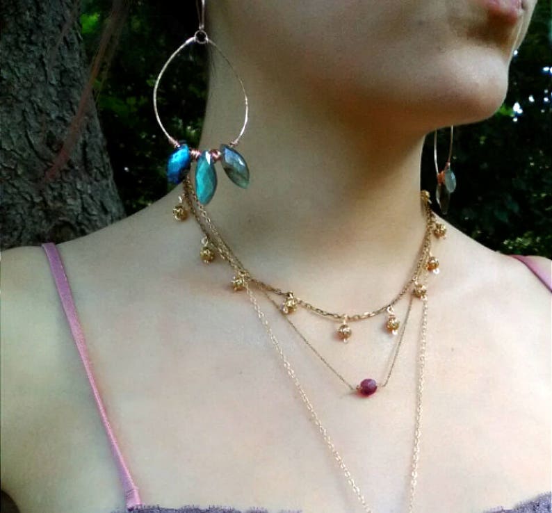 May include: Gold-toned jewelry set featuring large hoop earrings with blue-green gemstone accents and a layered necklace. The necklace includes small gold charms and a single, round, red gemstone pendant. The jewelry is worn against a light pink top.