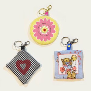 May include: Three fabric keychains with metal rings. One is round with a yellow and pink floral design. Another is a black and white checkered diamond shape with a red heart. The third is a blue square with a teddy bear illustration.