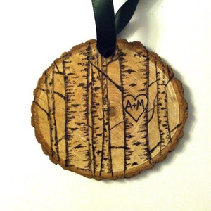 May include: A wooden ornament with a black ribbon, featuring a woodburned design of birch trees and a heart with the initials "A+M" inside.
