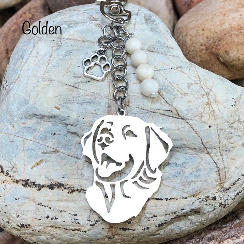 May include: A silver keychain featuring a cutout silhouette of a golden retriever dog's head. The keychain has a silver paw charm and white beads hanging from it. The text "Golden" is visible in the image.