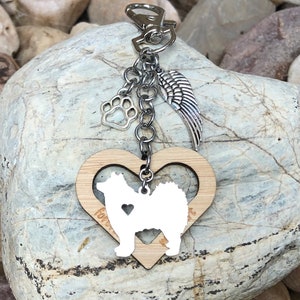 May include: Wooden heart-shaped keychain with a silhouette of a dog inside. The keychain has a silver chain with a paw print charm and a silver angel wing charm. The heart is engraved with the word "forever".