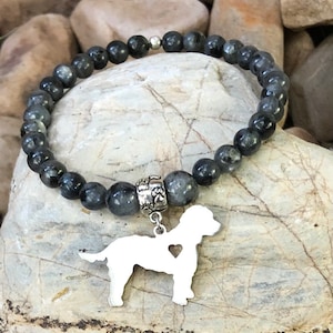 May include: A black beaded bracelet with a silver charm of a dog silhouette with a heart cutout. The bracelet is on a gray and brown rock background.