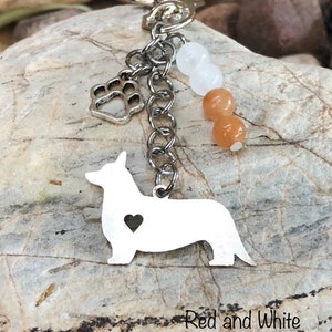 Puede incluir: Un llavero de plata con un dije en forma de silueta de corgi con un corazón recortado. El llavero también incluye un dije de pata de plata y tres cuentas: dos de color melocotón y una blanca.