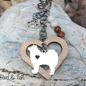 May include: A wooden heart-shaped keychain with a white silhouette of a dog inside. The keychain has a silver chain with a paw charm and red and tan beads.
