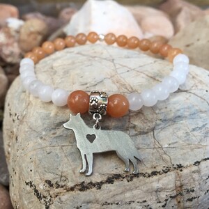 May include: A white and orange beaded bracelet with a silver dog charm. The charm has a heart cut out of it.
