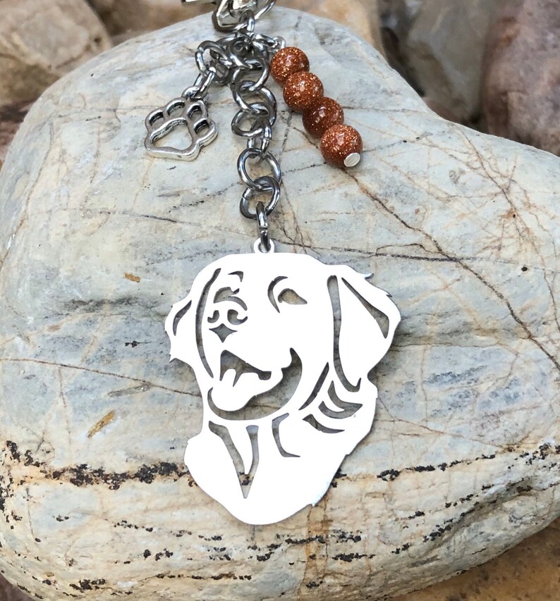 May include: Silver keychain featuring a cutout silhouette of a golden retriever dog's head. The keychain has a silver paw print charm and a chain with brown glitter beads.