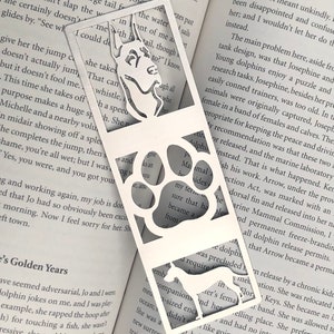 May include: A white metal bookmark with a cut-out design of a dog's head and a paw print. The bookmark is layered over a page of a book with text about a dog named Josephine.