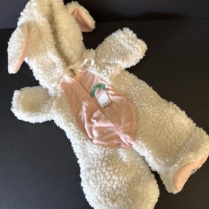 May include: A white lamb costume with pink accents. The costume features a hood with floppy ears, a pink bib with a lamb graphic, and attached sleeves and legs. The costume is made of a soft, textured material.