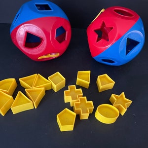 May include: A set of red and blue shape sorters with yellow plastic shapes. The sorters are shaped like balls with different cutouts for the shapes. The shapes include a star, a circle, a square, a triangle, and a rectangle.