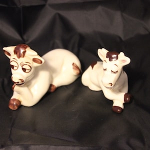 May include: Two ceramic cow figurines. The cows are white with brown spots and accents on their ears, hooves, and around their eyes. They are in a relaxed, reclining pose with their eyes closed. The background is black.