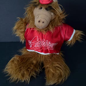 May include: A brown furry puppet with a red shirt that says "Alf" in white cursive. The puppet is wearing a red hat.