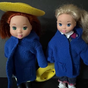 May include: Two dolls wearing blue coats and black boots. One doll has red hair and a yellow hat, the other has blonde hair. They are holding hands. The dolls have blue eyes and are facing the viewer.