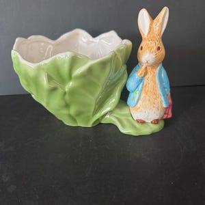 May include: A ceramic planter shaped like a green cabbage leaf, with a white interior. A small figurine of a rabbit wearing a blue jacket and pink bag stands beside the planter. The rabbit has brown fur and is in a standing position.