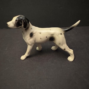 May include: A ceramic Dalmatian dog figurine. The dog is white with black spots and a black patch on its head. It is standing with its tail up, and its eyes are visible. The figurine is a decorative item.