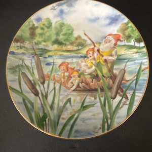 May include: A decorative plate featuring a whimsical scene of gnomes in a boat on a lake. The plate has a gold rim and depicts gnomes with red hats and yellow vests, surrounded by cattails and trees. The artwork is detailed and colorful.