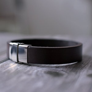 Pu&ograve; includere: Un braccialetto in pelle marrone con una chiusura in argento. Il braccialetto &egrave; appoggiato su una superficie in legno.