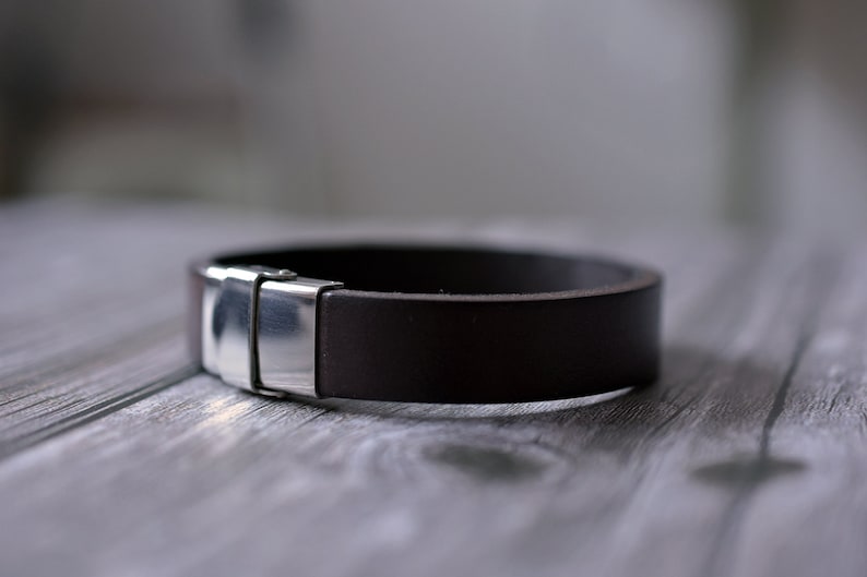 Pu&ograve; includere: Un braccialetto in pelle marrone con una chiusura in argento. Il braccialetto &egrave; appoggiato su una superficie in legno.