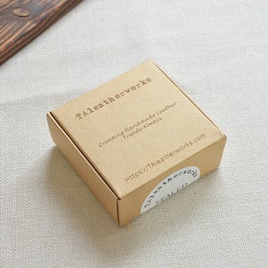 Pu&ograve; includere: Una scatola di cartone marrone con la scritta "Taleatherworks" stampata sulla parte superiore. La scatola ha un adesivo bianco con la scritta "Creating Handmade Leather Trends Always" e un indirizzo di sito web.