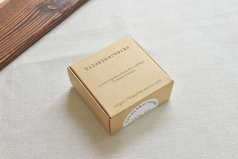 Pu&ograve; includere: Una scatola di cartone marrone con la scritta "Taleatherworks" stampata sulla parte superiore. La scatola ha un adesivo bianco con la scritta "Creating Handmade Leather Trends Always" e un indirizzo di sito web.