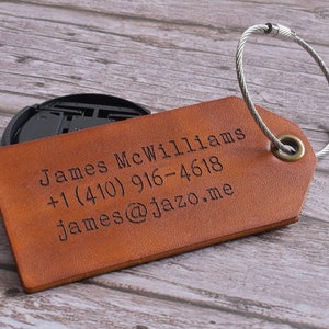 Może przedstawiać: Brązowa sk&oacute;rzana metka bagażowa z srebrnym metalowym oczkiem. Metka jest grawerowana tekstem "James McWilliams +1 (410) 916-4618 james@jazzo.me"