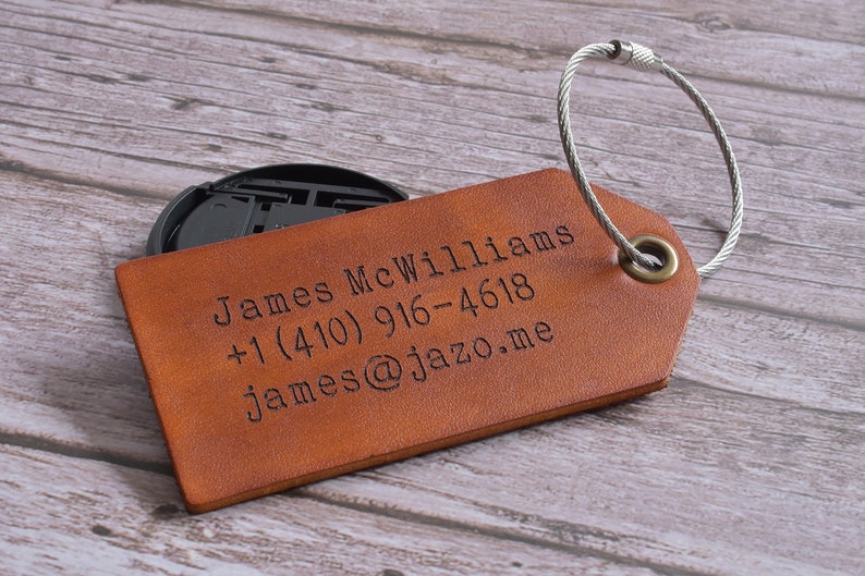 Może przedstawiać: Brązowa sk&oacute;rzana metka bagażowa z srebrnym metalowym oczkiem. Metka jest grawerowana tekstem "James McWilliams +1 (410) 916-4618 james@jazzo.me"