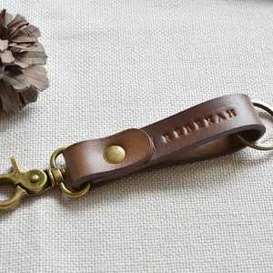 May include: A brown leather keychain with a brass key ring and a snap hook. The keychain is personalised with the name "REBEKAH" embossed in the leather.