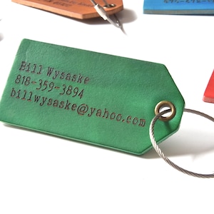 Może przedstawiać: Zielona sk&oacute;rzana metka bagażowa z metalowym k&oacute;łkiem i kablem. Metka ma wygrawerowany tekst "Bill Wysaske, 818-359-3894, billwysaske@yahoo.com".