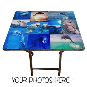 May include: A folding table with a blue and white dolphin themed tabletop. The table is made of wood and has a brown finish. The tabletop is made of a durable material and features a collage of dolphin images.