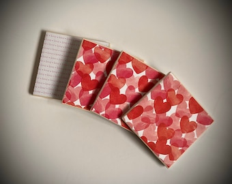 May include: Four square coasters with a white background and a pattern of red watercolor hearts. The text "love love love love" is printed on one coaster.