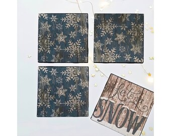 Może przedstawiać: Zestaw czterech niebieskich i złotych podkładek pod kubki z płatkami śniegu i drewnianym znakiem z napisem "Let it Snow".