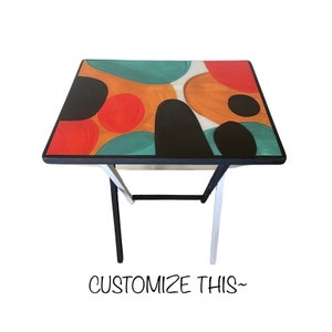 May include: A black and white metal folding table with a colorful abstract design on the tabletop. The design features circles, ovals, and abstract shapes in shades of red, orange, yellow, green, and blue.