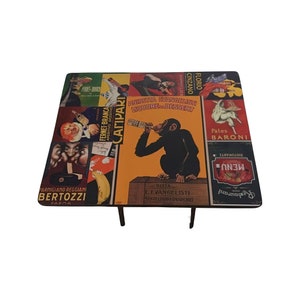 May include: A square table with a colorful, vintage-style design featuring various Italian brand logos and advertisements. The table top is made of wood and has a glossy finish. The table has four black legs.
