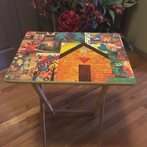 May include: A wooden folding table with a colorful painted top. The table top features a collage of images and text, including the words "Cherish Family", "Share", "Grow", "Love Life", "Take Good Care", "Remember", "Don't Grow Too Fast", "Make Music", "Garden", "Laugh Cry", "Just Play", "Together", "Celebrate", "Plan a Vacation", "Be Creative", "Have Fun Holidays", "Watch Movies", "Love Cow", "Be Grateful", and "Follow Your Heart".