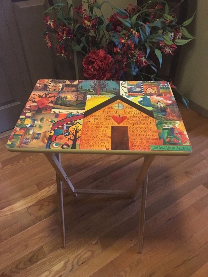 May include: A wooden folding table with a colorful painted top. The table top features a collage of images and text, including the words "Cherish Family", "Share", "Grow", "Love Life", "Take Good Care", "Remember", "Don't Grow Too Fast", "Make Music", "Garden", "Laugh Cry", "Just Play", "Together", "Celebrate", "Plan a Vacation", "Be Creative", "Have Fun Holidays", "Watch Movies", "Love Cow", "Be Grateful", and "Follow Your Heart".