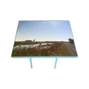 May include: A light blue table with a rectangular glass top featuring a photograph of a grassy field with a body of water in the background. The sun is shining brightly in the sky.