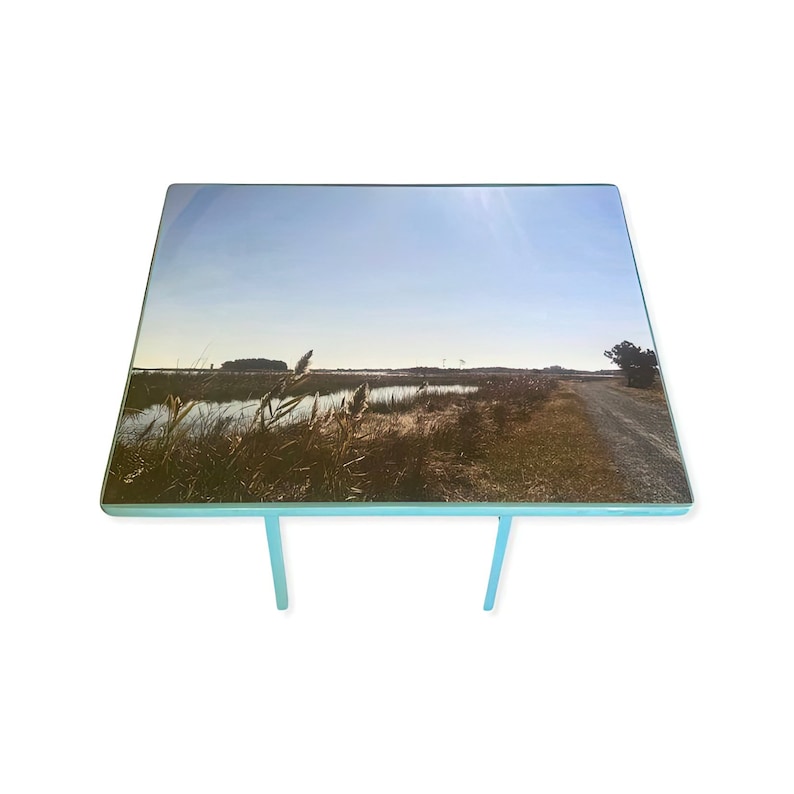 May include: A light blue table with a rectangular glass top featuring a photograph of a grassy field with a body of water in the background. The sun is shining brightly in the sky.