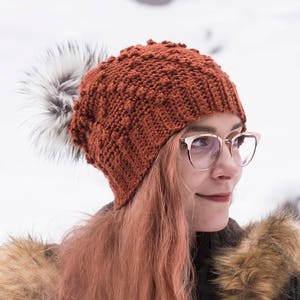 May include: A woman wearing a rust-colored crocheted beanie with a white pom pom and clear framed glasses. She is wearing a green jacket with a fur-lined hood.