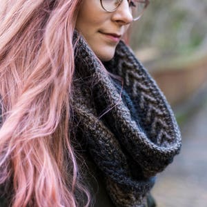 Könnte beinhalten: Eine Frau mit pinken Haaren trägt einen grauen und blauen Strick Schal und eine Brille. Sie schaut nach unten und ihre Haare bedecken den größten Teil ihres Gesichts.