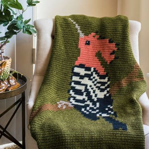 May include: A green crocheted throw blanket featuring a colourful woodpecker design. The blanket is draped over a white armchair.
