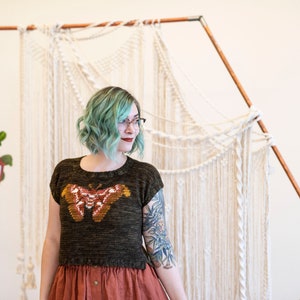 May include: A woman with teal blue hair wearing a dark green knitted top with a brown and orange butterfly design. She is standing in front of a white macrame wall hanging.