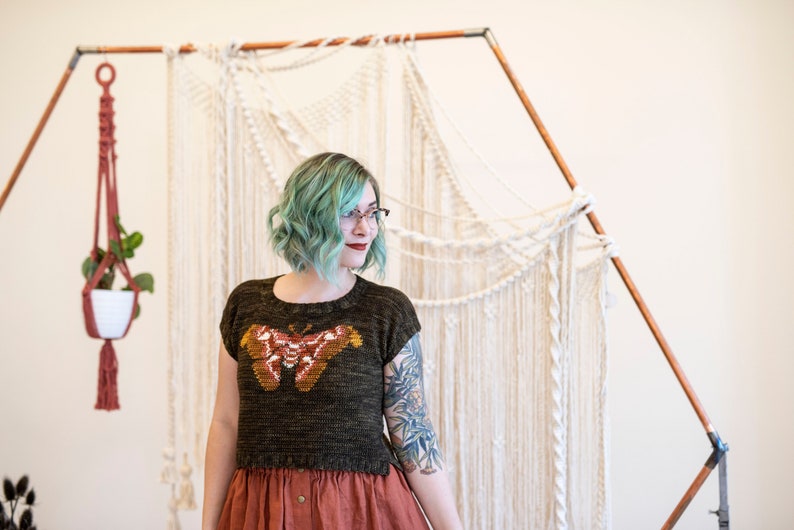 May include: A woman with teal blue hair wearing a dark green knitted top with a brown and orange butterfly design. She is standing in front of a white macrame wall hanging.