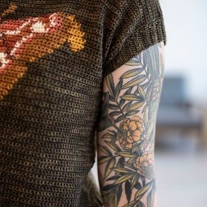 May include: A person wearing a brown knit shirt with a colorful design and a floral tattoo on their arm.