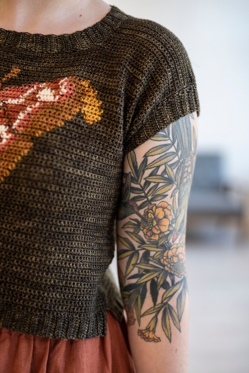 May include: A person wearing a brown knit shirt with a colorful design and a floral tattoo on their arm.