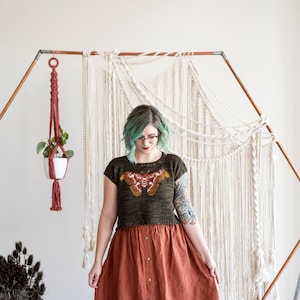 May include: A woman wearing a green and brown knit top with a butterfly design, a rust-colored skirt, and glasses. She is standing in front of a white macrame wall hanging.
