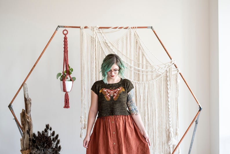 May include: A woman wearing a green and brown knit top with a butterfly design, a rust-colored skirt, and glasses. She is standing in front of a white macrame wall hanging.